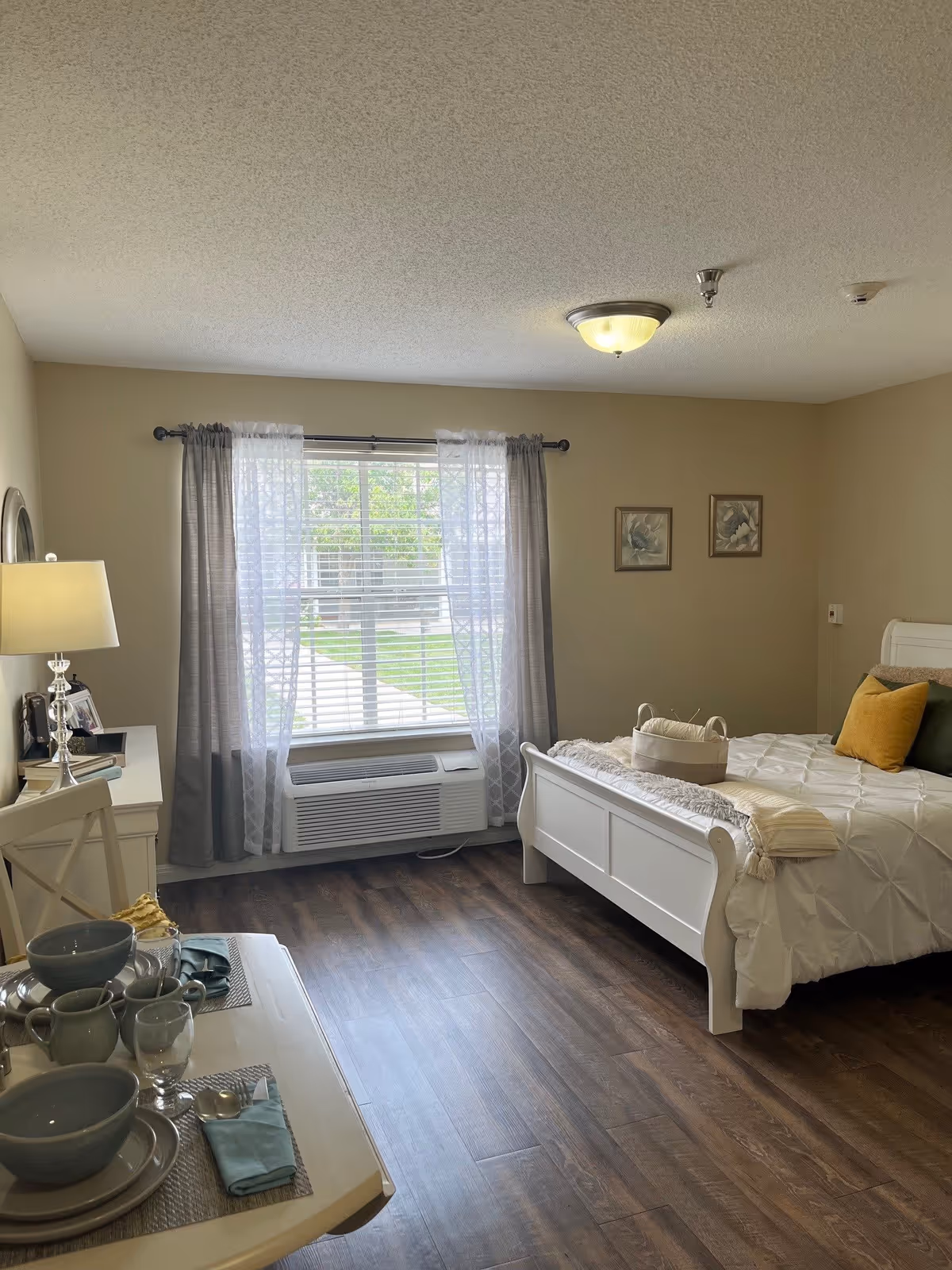 A bright bedroom with a white bed featuring a white comforter and yellow and green pillows. There is a window with sheer and gray curtains letting in natural light. A white desk with a lamp and framed photos is on the left side, and a small dining table set with bowls, plates, glasses, and napkins is in the foreground. The floor is dark wood, and two framed floral pictures hang on the wall.