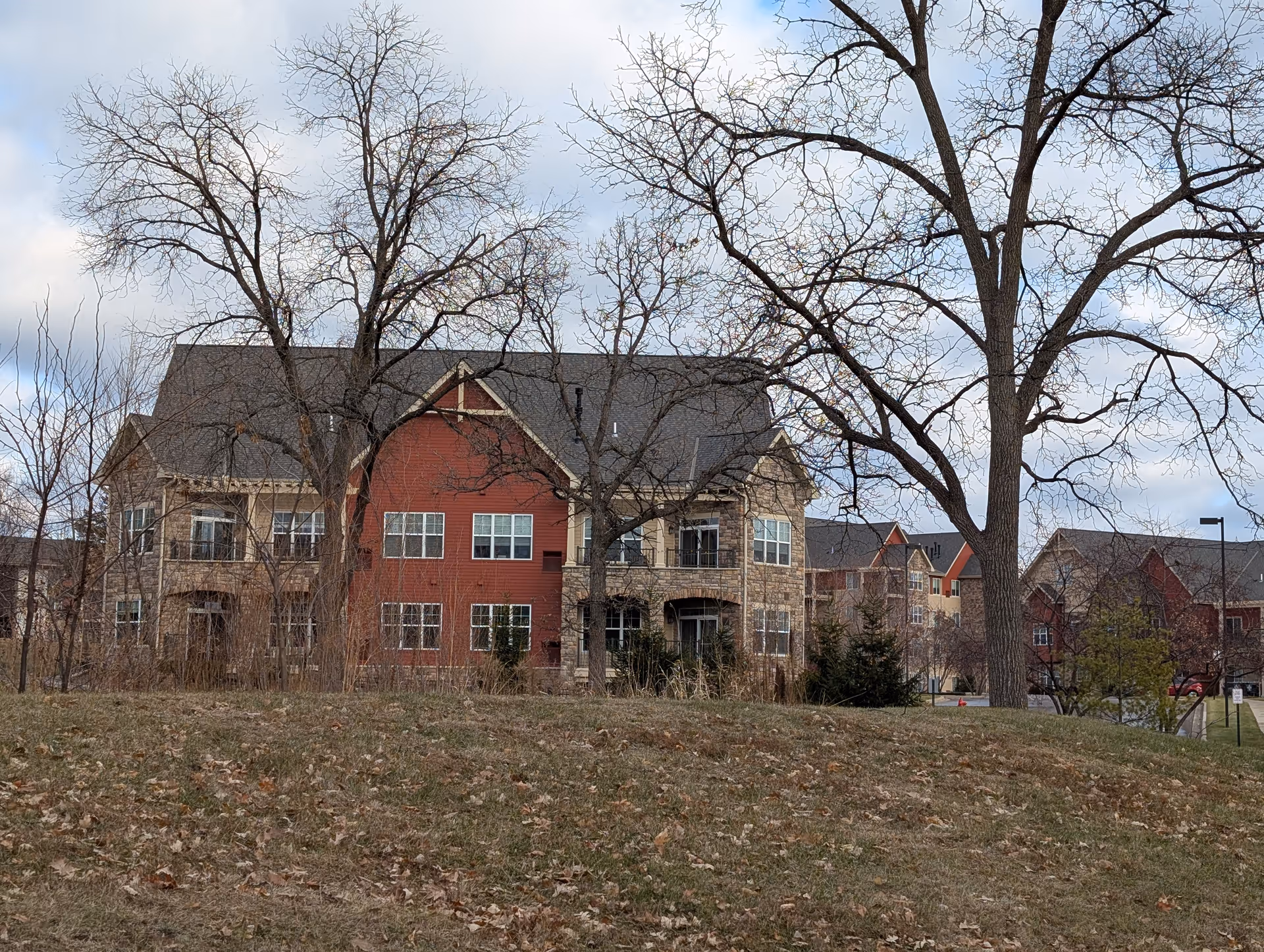 Front view of a multi-story red and stone apartment building with leafless trees and a grassy foreground.