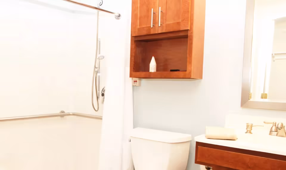 A bright bathroom featuring a white toilet, a wooden cabinet mounted on the wall above the toilet, a shower area with a white curtain and a handheld showerhead, and a vanity with a sink, faucet, and a folded towel. A mirror is partially visible on the right side.