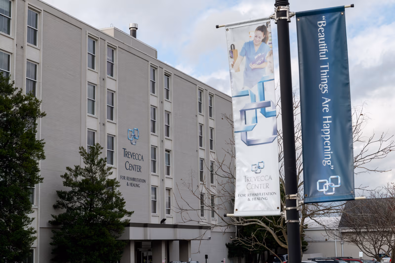 Exterior view of the Trevecca Center for Rehabilitation and Healing building with multiple windows and a sign on the wall displaying the facility's name. Two vertical banners hang from a pole in the foreground, one showing a healthcare worker and the other with the text 'Beautiful Things Are Happening'. Trees and a cloudy sky are also visible.