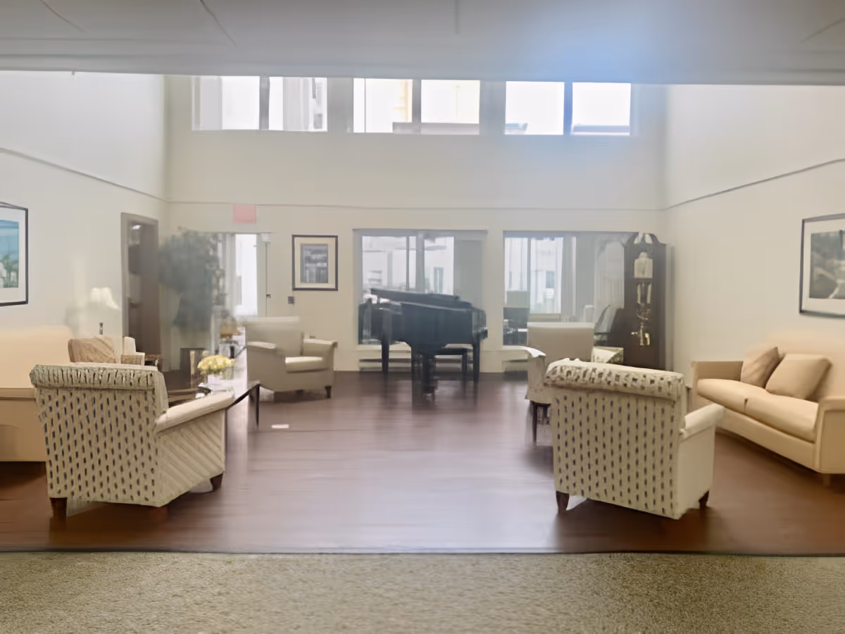 A spacious living room area with several upholstered armchairs and sofas arranged around a wooden floor. A grand piano is positioned near the back wall, which features large windows letting in natural light. There is a tall grandfather clock to the right and framed artwork on the walls.