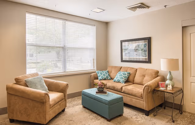 A cozy living room with a tan sofa and matching armchair, both adorned with blue patterned throw pillows. A teal ottoman sits in front of the sofa with a small plant and book on top. A side table with a lamp and decorative items is next to the sofa. A large window with blinds lets in natural light, and a framed picture hangs on the wall above the sofa.