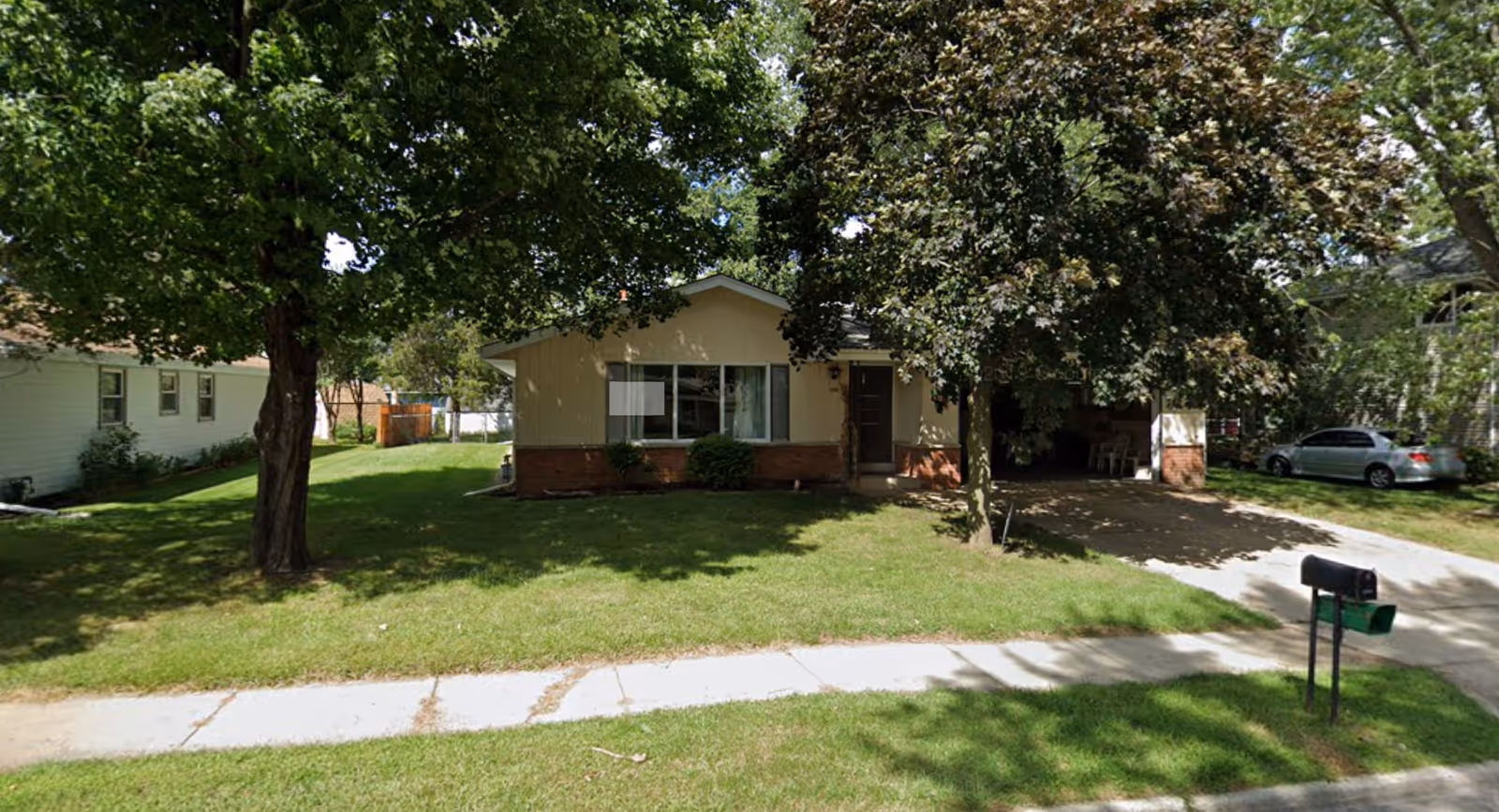 Single-story house with a beige exterior and brick accents, surrounded by green grass and large trees. A driveway leads to a carport on the right side of the house, and a sidewalk runs along the front lawn. A mailbox is visible near the sidewalk.