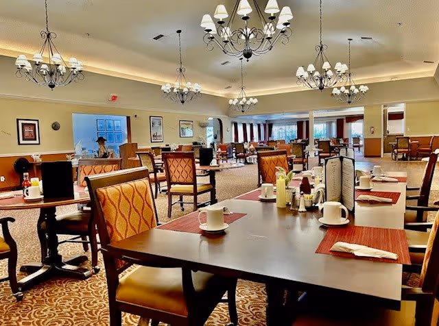 A spacious dining room with multiple tables and chairs arranged neatly. Each table is set with white coffee cups, napkins, menus, and condiments. The room features elegant chandeliers hanging from the ceiling, patterned carpet flooring, and framed artwork on the walls. Large windows with curtains allow natural light into the room.
