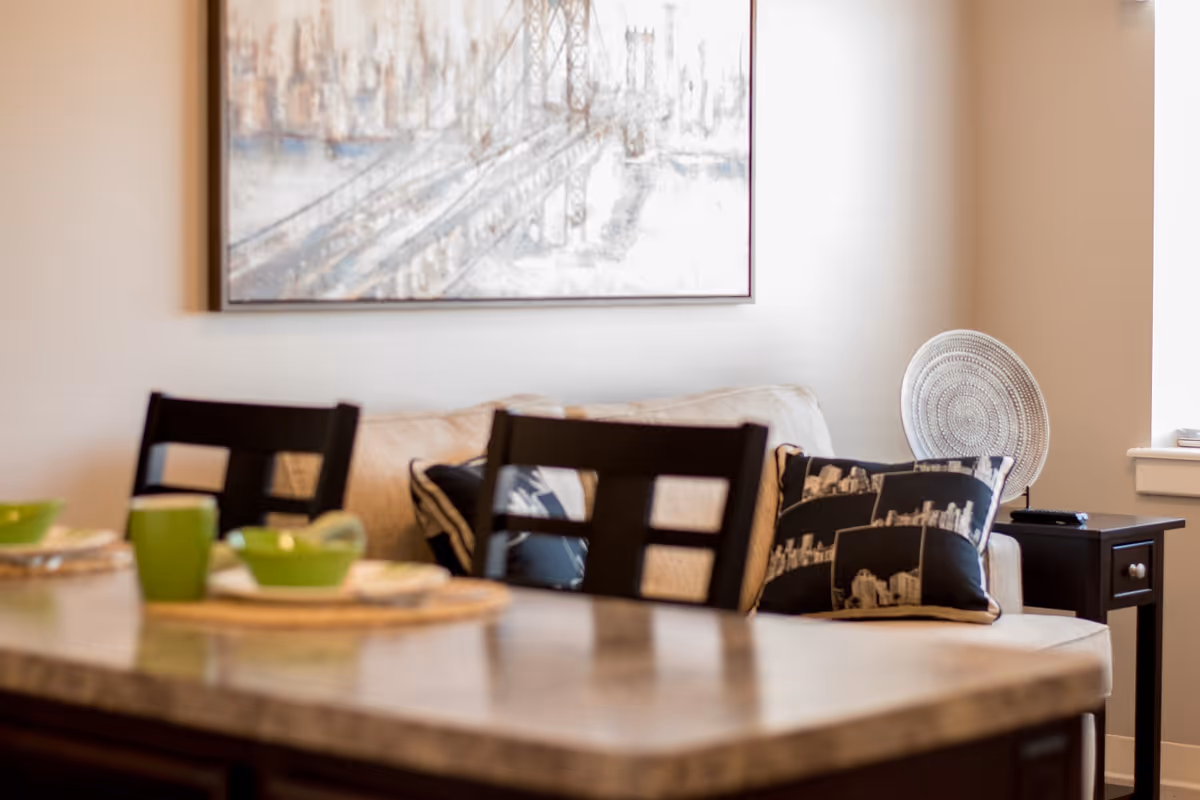 A cozy living area with a beige couch adorned with black and white cityscape pillows, a dark wooden side table with a decorative circular plate, and a large framed artwork of a bridge on the wall. In the foreground, there is a marble countertop with green bowls and cups placed on woven placemats, along with two dark wooden chairs.