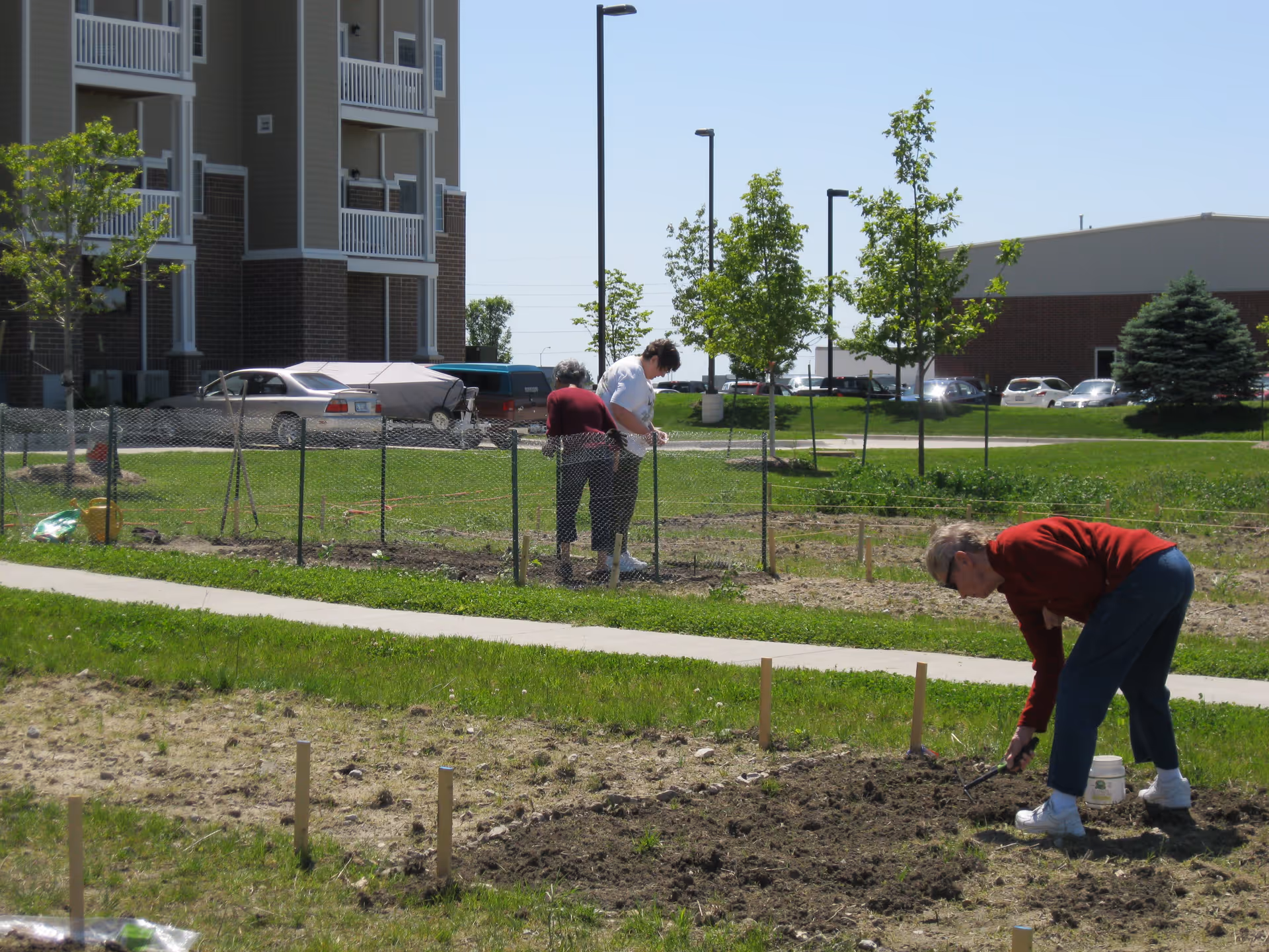Three elderly people gardening outdoors near a residential building. Two individuals are working inside a fenced garden area, while another person is tending to soil outside the fence. There are young trees, a sidewalk, parked cars, and a building in the background under a clear blue sky.