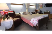 A cozy bedroom in a senior living facility featuring a hospital-style bed with pink and white bedding, a brown recliner chair, a side table with a lamp and a vase of flowers, and a television mounted on the wall. The room has light blue walls and a window with a curtain.