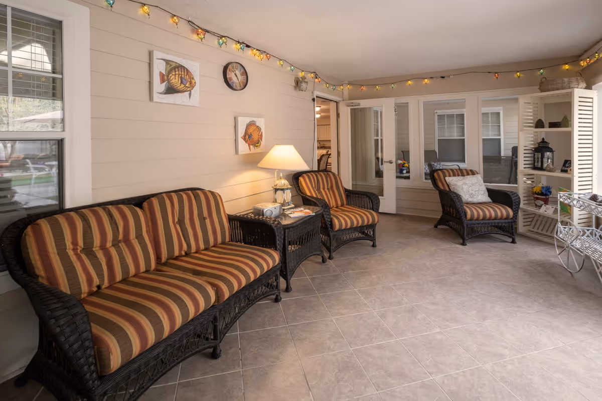 A cozy indoor seating area with a striped cushioned wicker sofa and two matching armchairs. The room has beige tiled flooring, light-colored walls decorated with fish artwork and a clock, and string lights hanging along the ceiling. A small table with a lamp and a radio sits between the sofa and one armchair, and a white shelving unit with decorative items is visible in the corner.