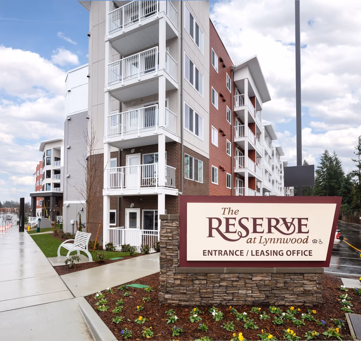 Exterior view of The Reserve at Lynnwood building with multiple balconies, a sidewalk, a bench, landscaped flower beds, and a large sign indicating the entrance and leasing office.