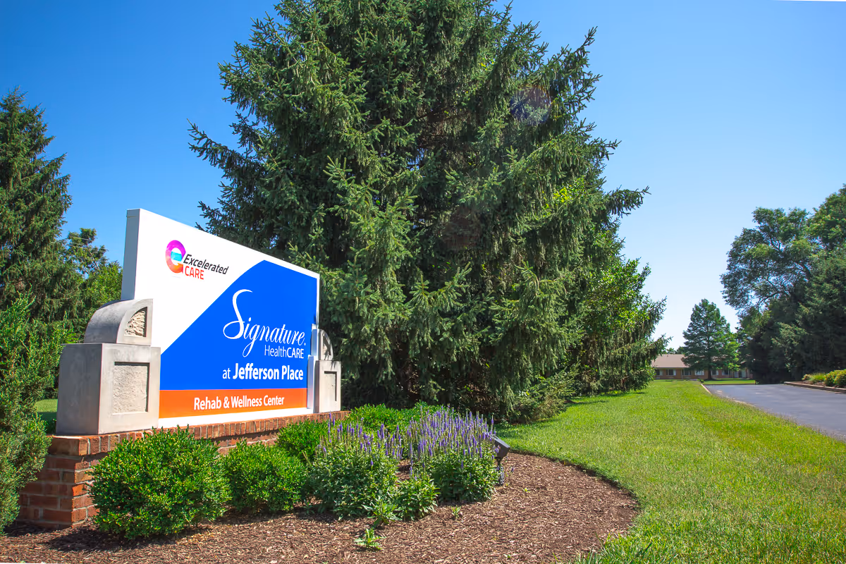 Outdoor view of the entrance sign for Signature HealthCARE at Jefferson Place Rehab & Wellness Center, surrounded by green bushes, purple flowers, and tall pine trees under a clear blue sky.