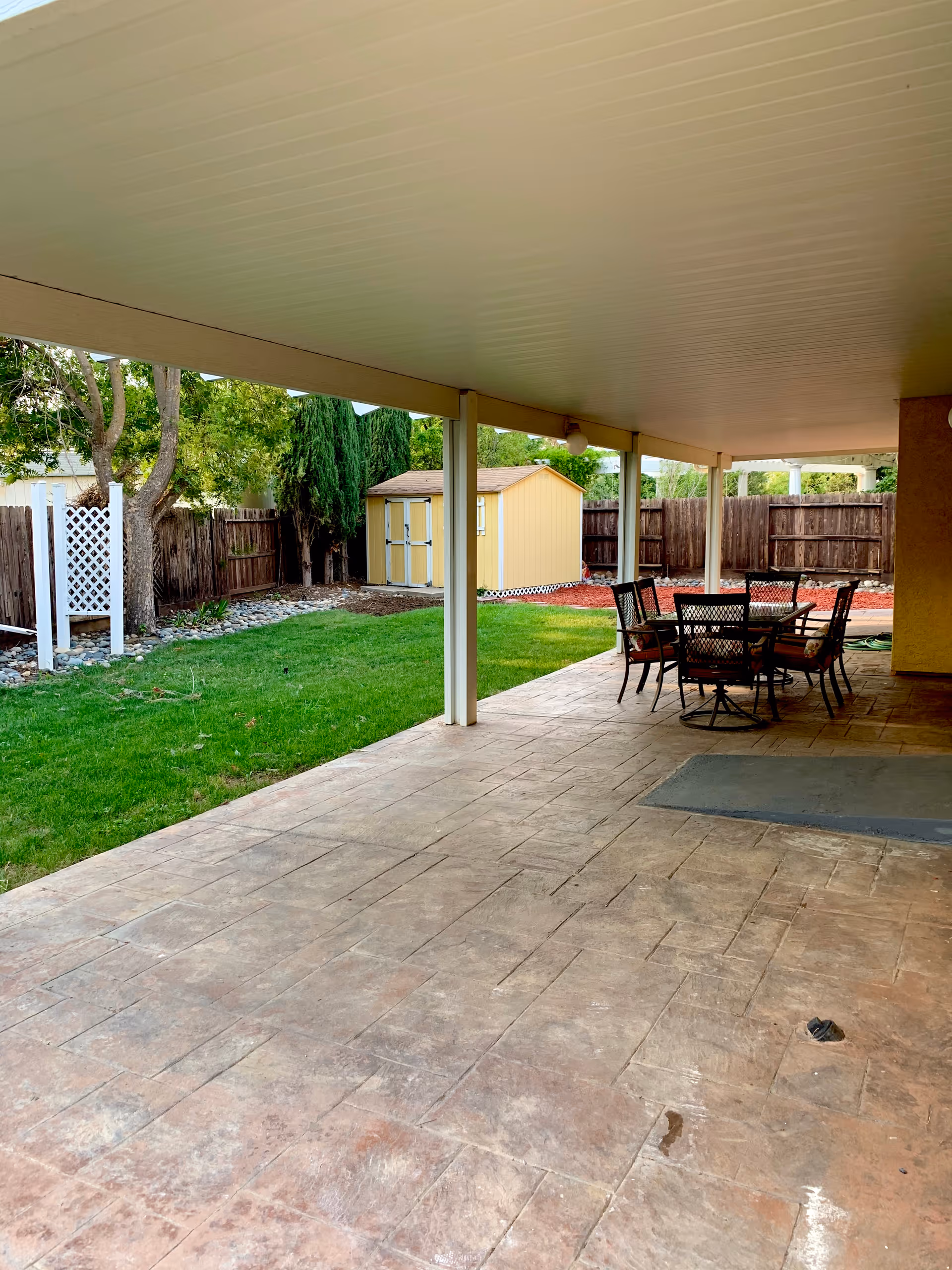 Covered backyard patio with an outdoor dining table, green lawn, and a yellow storage shed.