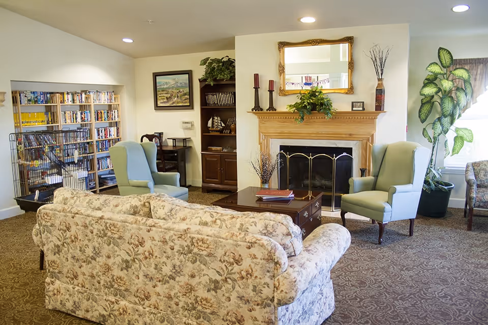 Cozy senior living common room with a floral sofa, two wingback chairs, a fireplace, bookshelves and indoor plants.