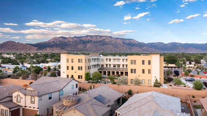 Aerial view of The Watermark at Cherry Hills facility with a three-story beige building surrounded by residential houses, trees, and a clear sky with mountains in the background.