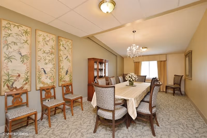 A formal dining room with a long rectangular table covered with a beige tablecloth and surrounded by eight upholstered wooden chairs. A crystal chandelier hangs above the table. On the left wall, there are three framed paintings featuring birds and plants. A wooden china cabinet with glass doors displaying plates is positioned against the far wall. The room has beige walls, a window with beige curtains, and patterned carpet flooring.