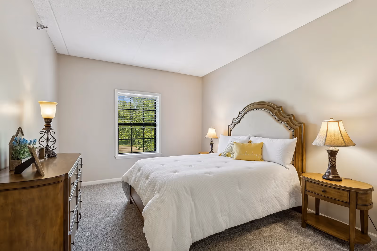 A bedroom with a large bed featuring a decorative wooden headboard, white bedding, and yellow accent pillows. There are two wooden nightstands with lamps on either side of the bed. A wooden dresser with decorative items and a lamp is positioned against the opposite wall. A window with black blinds shows green trees outside. The walls are painted beige and the floor is carpeted.
