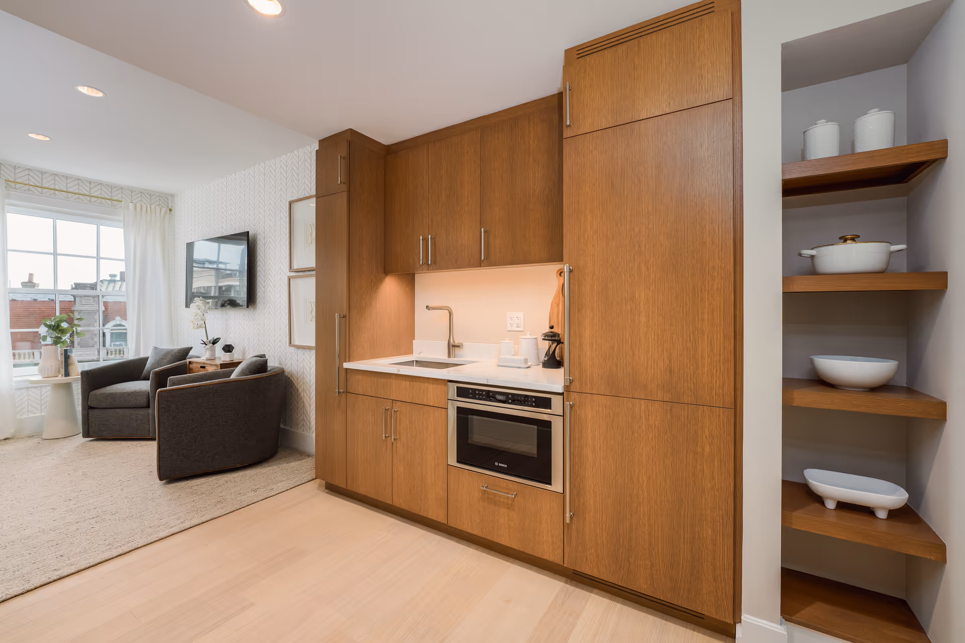 Modern kitchenette with wooden cabinets, a built-in microwave, a sink, and open shelves holding white dishes. Adjacent to the kitchenette is a cozy sitting area with two gray armchairs, a small side table with a plant, and a wall-mounted TV. Large window with sheer white curtains allows natural light into the space.