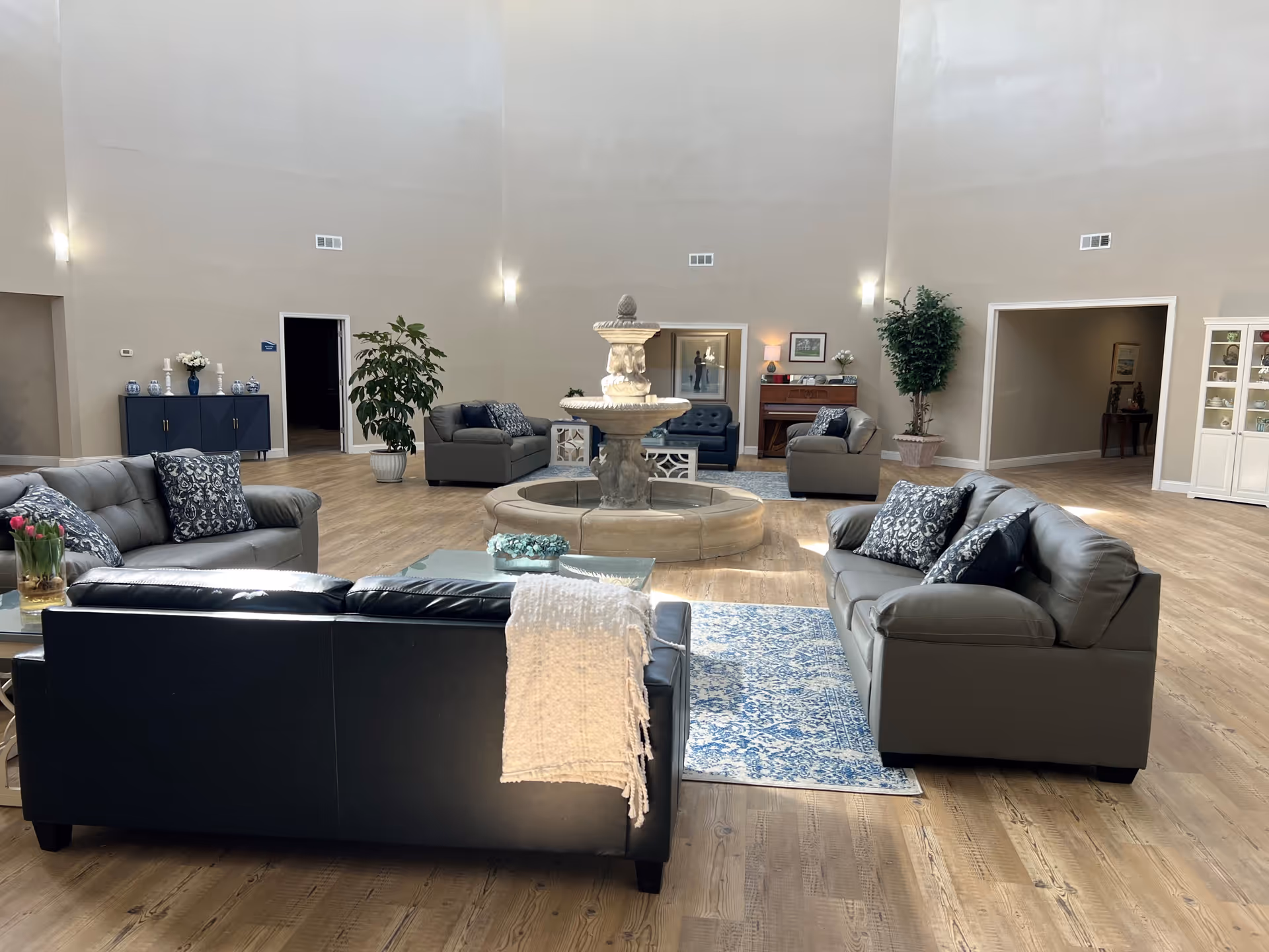 A spacious senior living facility common area with multiple gray sofas arranged around a central stone fountain. The room has high ceilings, light-colored walls, wooden flooring, and decorative plants. There are blue patterned pillows on the sofas and a blue and white rug on the floor. In the background, there is a piano, a cabinet with decorative items, and open doorways leading to other rooms.