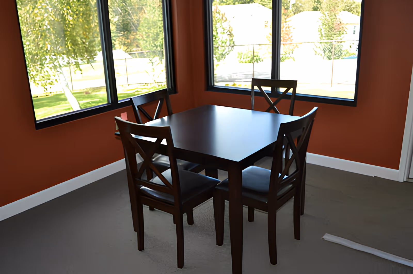 A small dining area with a square dark wooden table and four matching chairs with cushioned seats, situated near two large windows with a view of green trees and a fenced yard outside. The walls are painted a warm reddish-brown color and the floor is carpeted.