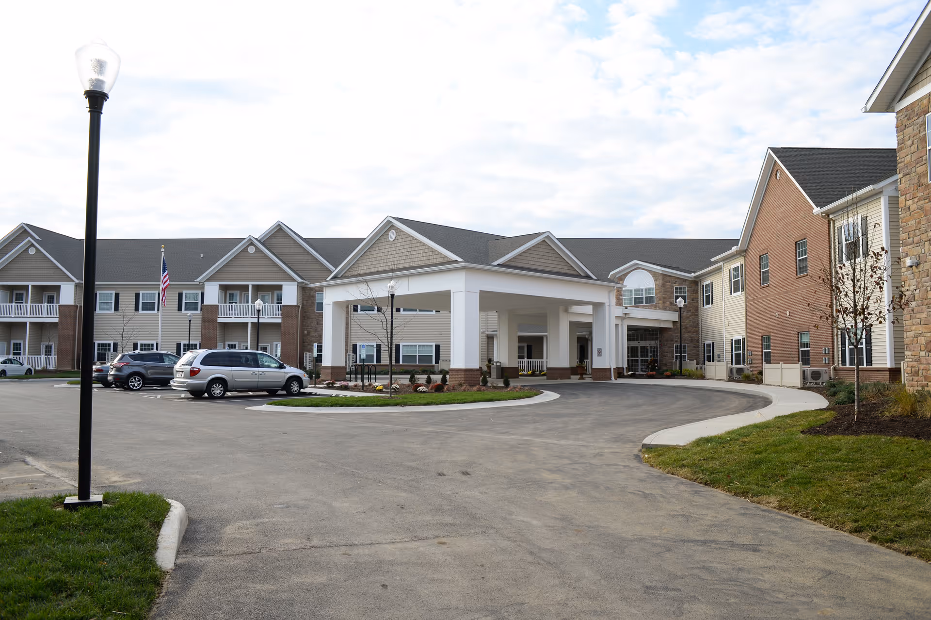 Exterior view of a senior living facility named Danbury Tallmadge showing a large building with multiple windows, a covered entrance with white pillars, a driveway, parked cars, a flagpole with an American flag, and some landscaping with grass and small trees.
