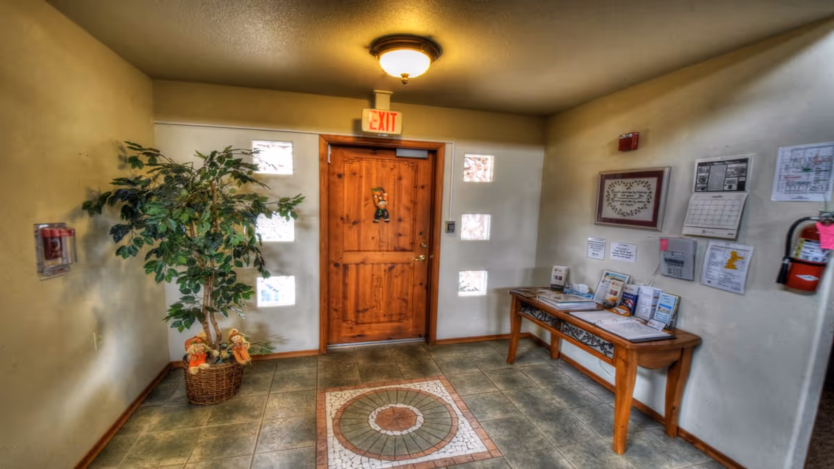 Interior view of a small entryway or lobby area with a wooden door featuring a decorative hanging. The door is surrounded by six small square windows. To the left, there is a potted artificial plant with two small scarecrow decorations at its base. On the right side, a wooden table holds various brochures and papers, with a calendar and framed artwork hanging on the wall above it. A fire extinguisher is mounted on the far right wall, and an exit sign is illuminated above the door.