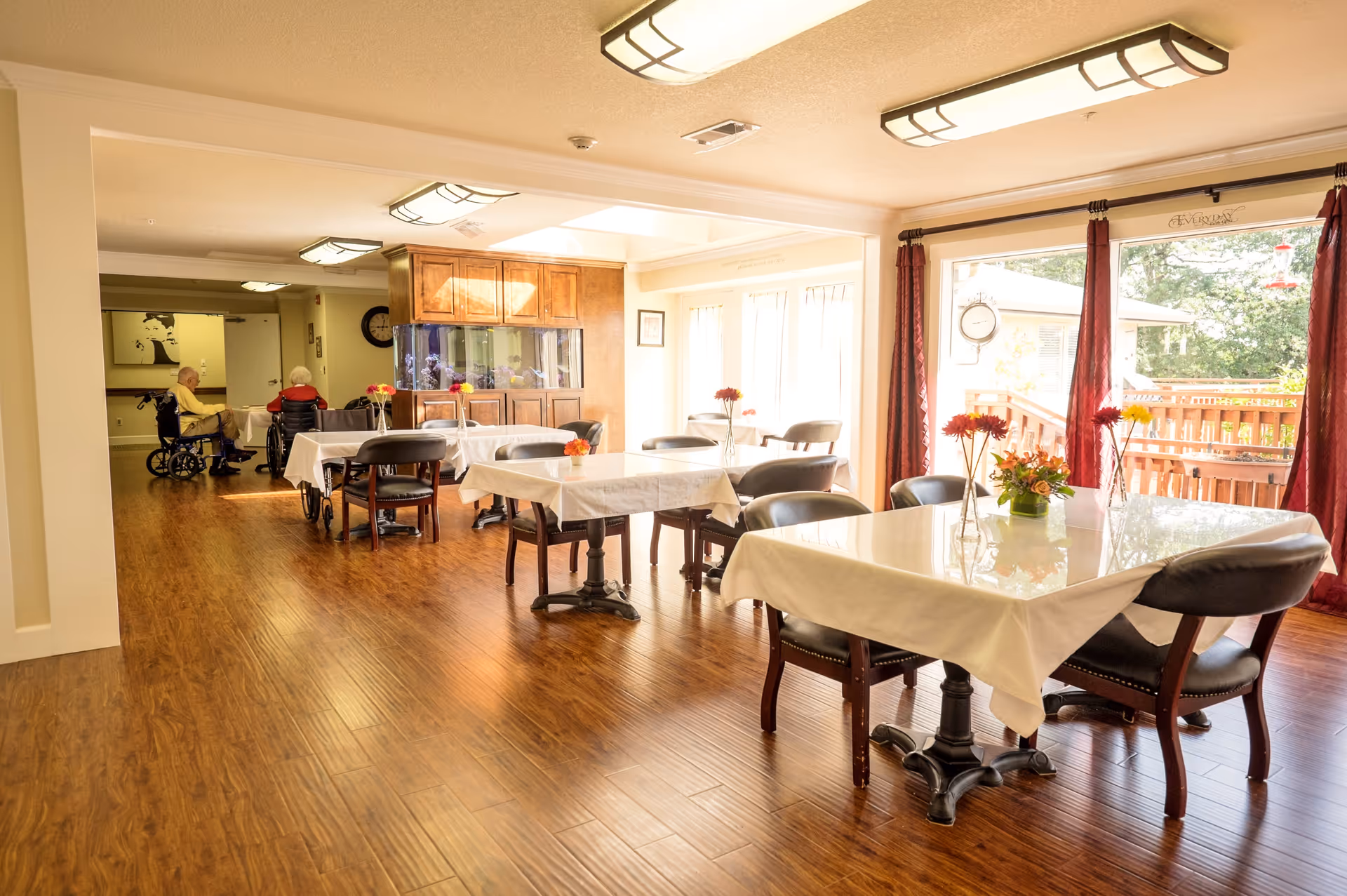 Well-lit senior living dining room with tables covered in white tablecloths, chairs and flower centerpieces, hardwood floors, and two people in wheelchairs near the back.