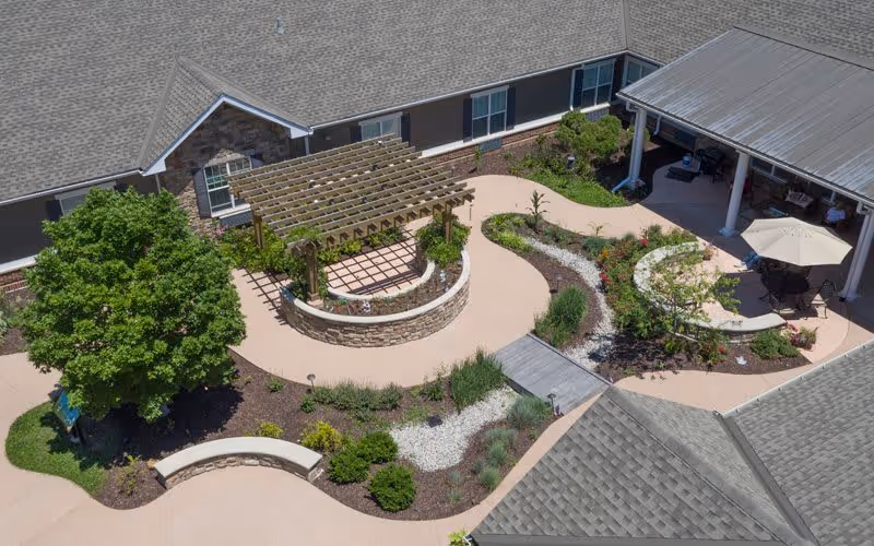 Aerial view of a landscaped outdoor courtyard at a senior living facility with a wooden pergola, curved stone seating walls, walking paths, various plants and shrubs, a large tree, and a covered patio area with tables and chairs under an umbrella.