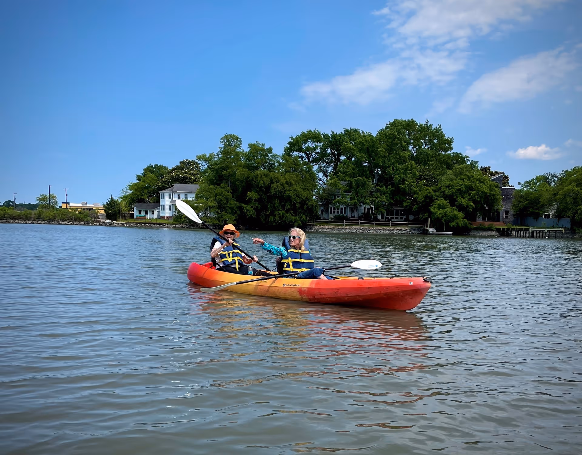 Two elderly women wearing life jackets are kayaking on a calm body of water with trees and houses visible in the background under a partly cloudy blue sky.
