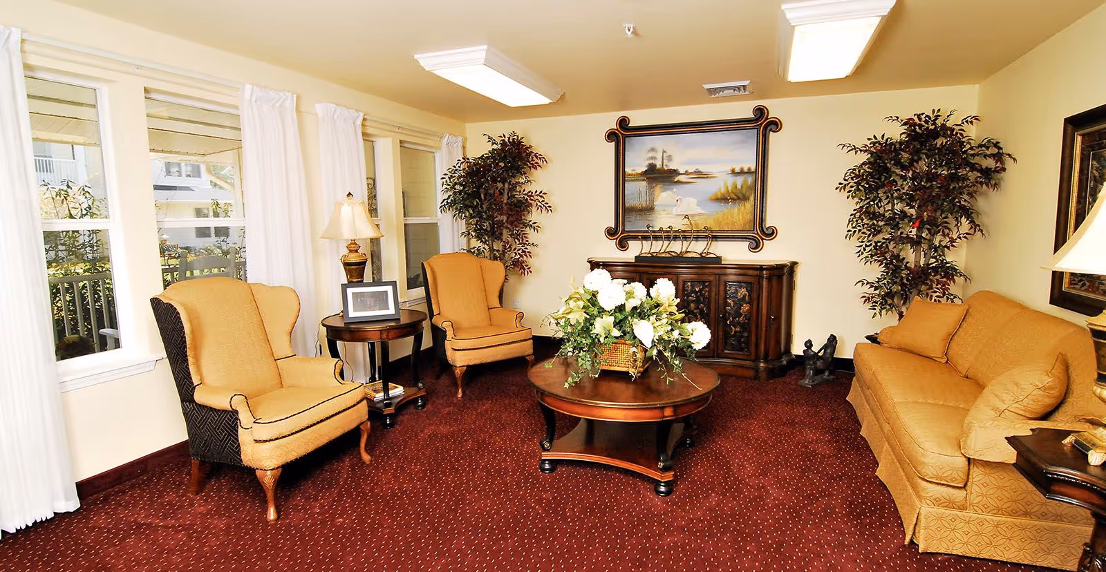 Bright, traditionally furnished living room with upholstered armchairs and a sofa arranged around a round coffee table topped with a floral arrangement.