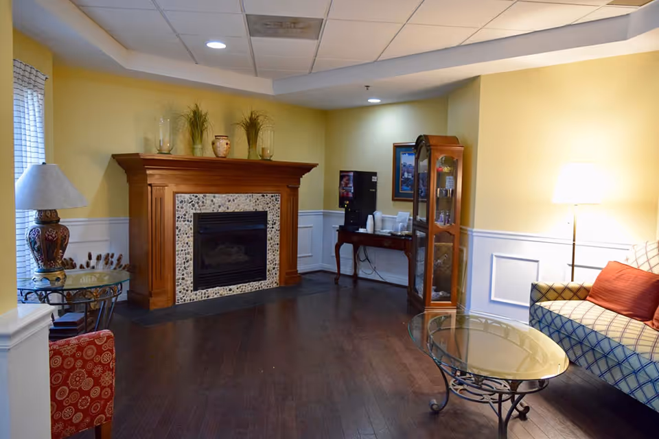 A cozy living room area with yellow walls and dark wooden flooring featuring a fireplace with a wooden mantel decorated with vases and plants. To the right, there is a glass coffee table in front of a patterned sofa with red cushions and a floor lamp. On the left side, there is a small round glass side table with a decorative lamp and a red patterned chair. In the back corner, a small table holds a coffee machine and cups, next to a wooden display cabinet with glass doors and a framed picture on the wall.