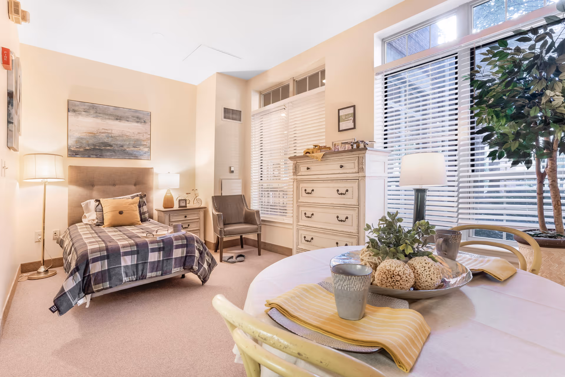 A cozy bedroom in a senior living facility featuring a single bed with plaid bedding and pillows, a nightstand with a lamp and clock, a comfortable chair, a tall white dresser, and a round table set with plates, cups, and decorative items. Large windows with white blinds allow natural light to fill the room, and a potted plant adds greenery to the space.