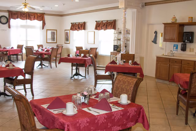 Dining room with multiple tables covered in red tablecloths, each set with folded napkins, cups, and utensils. The room has tiled floors, large windows with brown valances, and wooden chairs with patterned upholstery. There is a small kitchenette area with cabinets, a microwave, and a sink in the background.