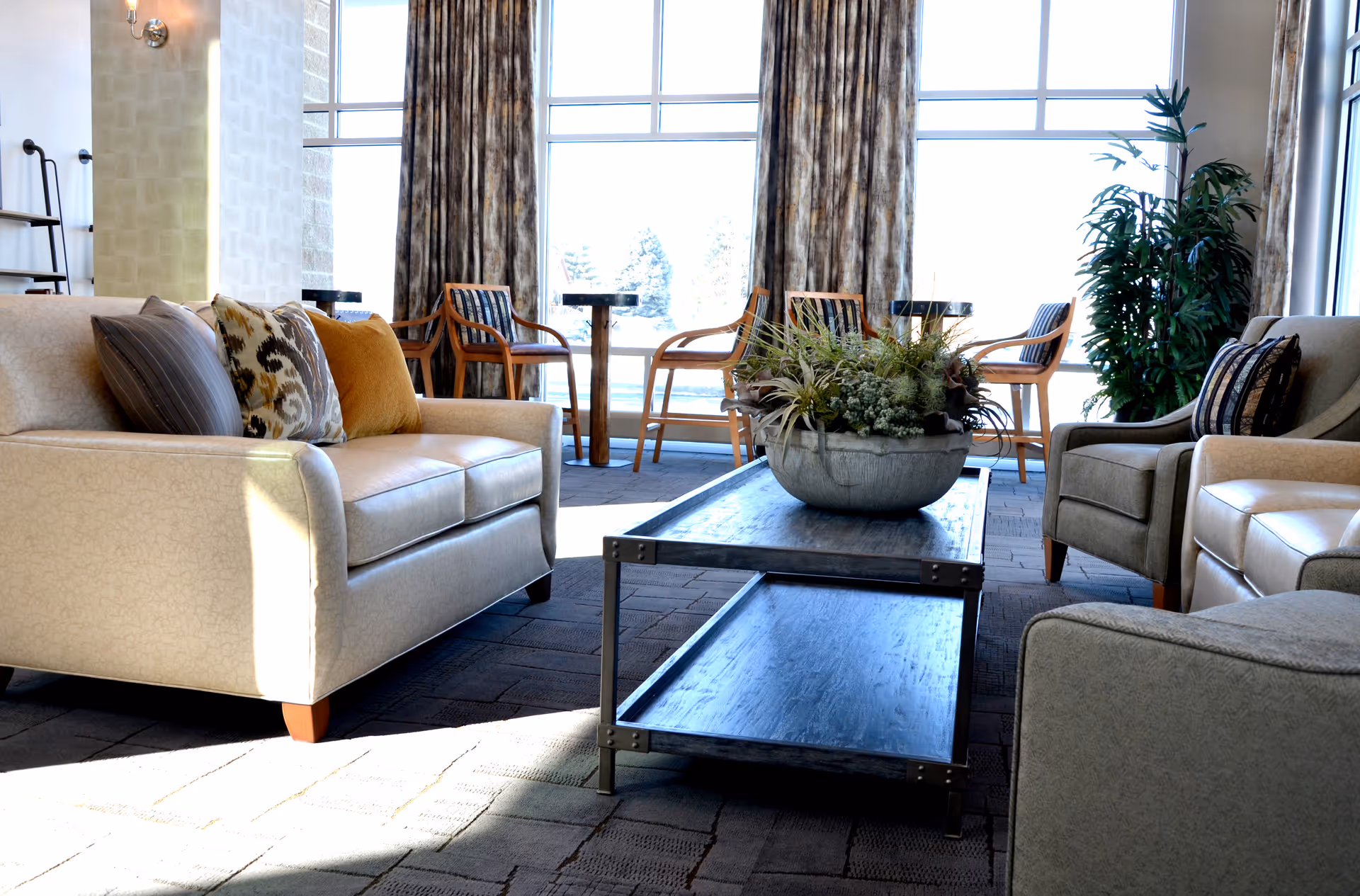 A bright and cozy living room area with beige sofas and armchairs arranged around a rectangular wooden coffee table with a large decorative plant centerpiece. Behind the seating area, there are large windows with patterned curtains and wooden chairs with small round tables near the windows. A tall green plant is placed near the window on the right side.