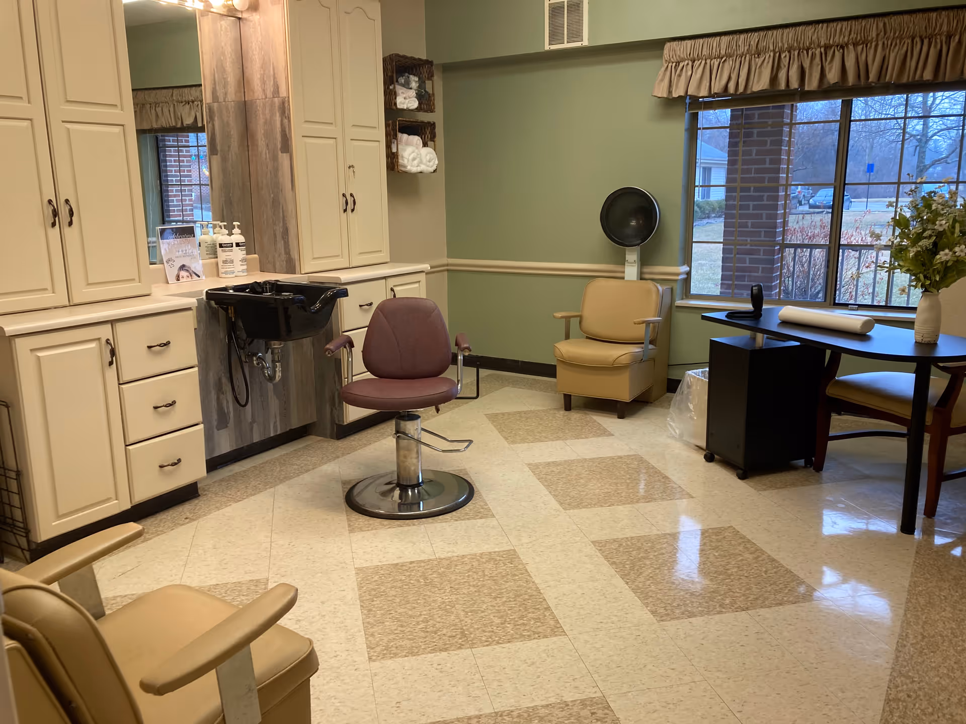 Interior view of a hair salon area in an assisted living facility with a salon chair in front of a black hair washing sink, beige cabinets, a beige armchair with a hair dryer hood, a table with a vase of flowers, and a large window showing an outdoor view.