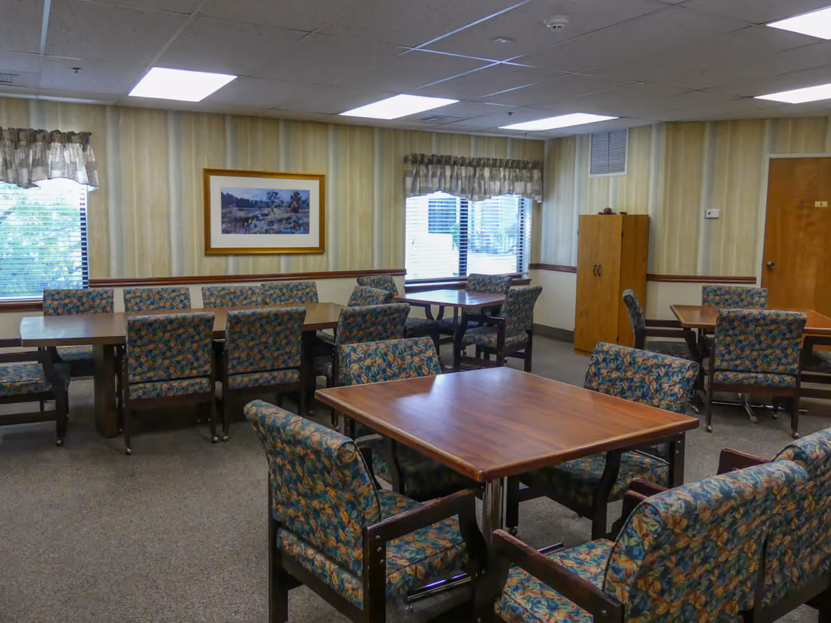 A dining room in a senior living facility with multiple wooden tables and floral upholstered chairs. The room has beige striped wallpaper, two windows with valances, a framed landscape painting on the wall, and a wooden cabinet near a closed door.