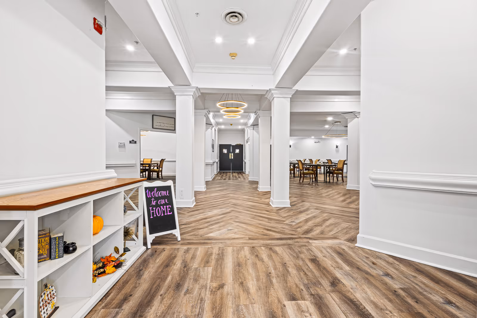 Bright open hallway leading to a communal dining area with wood floors, white columns, and a small sign that says "Welcome to our HOME".