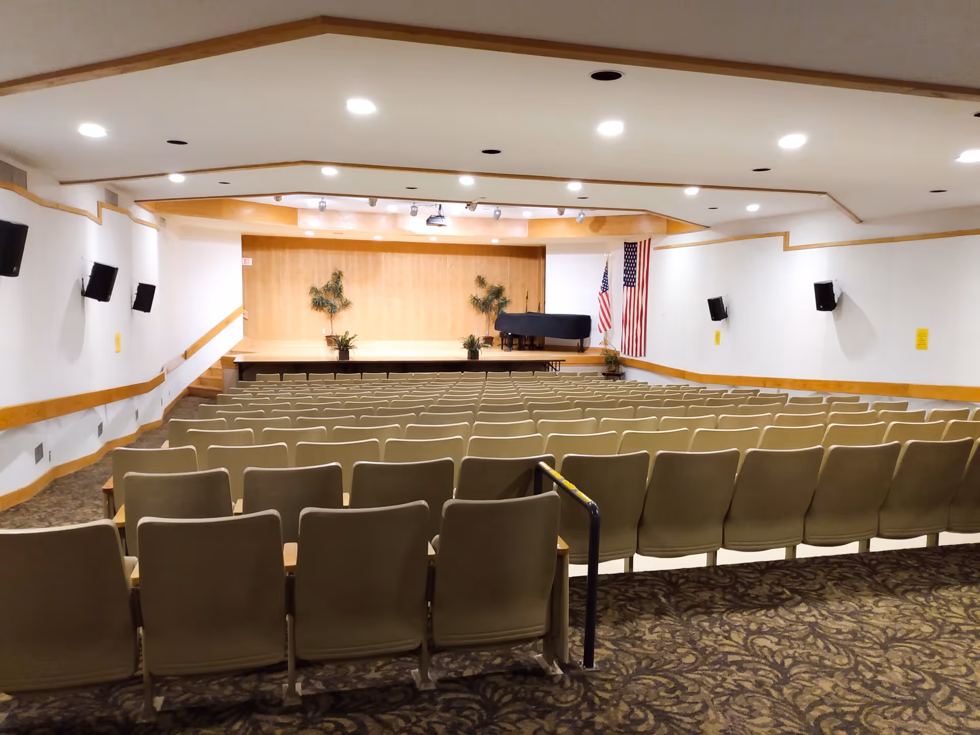 Empty auditorium with rows of beige chairs facing a stage with a piano and American flags.