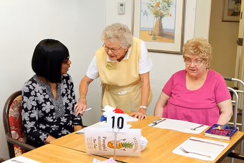 Three women sitting and standing around a dining table in a senior living facility. One woman in a yellow apron is standing and engaging with two seated women, one wearing a black and white patterned top and the other in a pink shirt. The table has place settings, a menu, and a table number 10 sign.