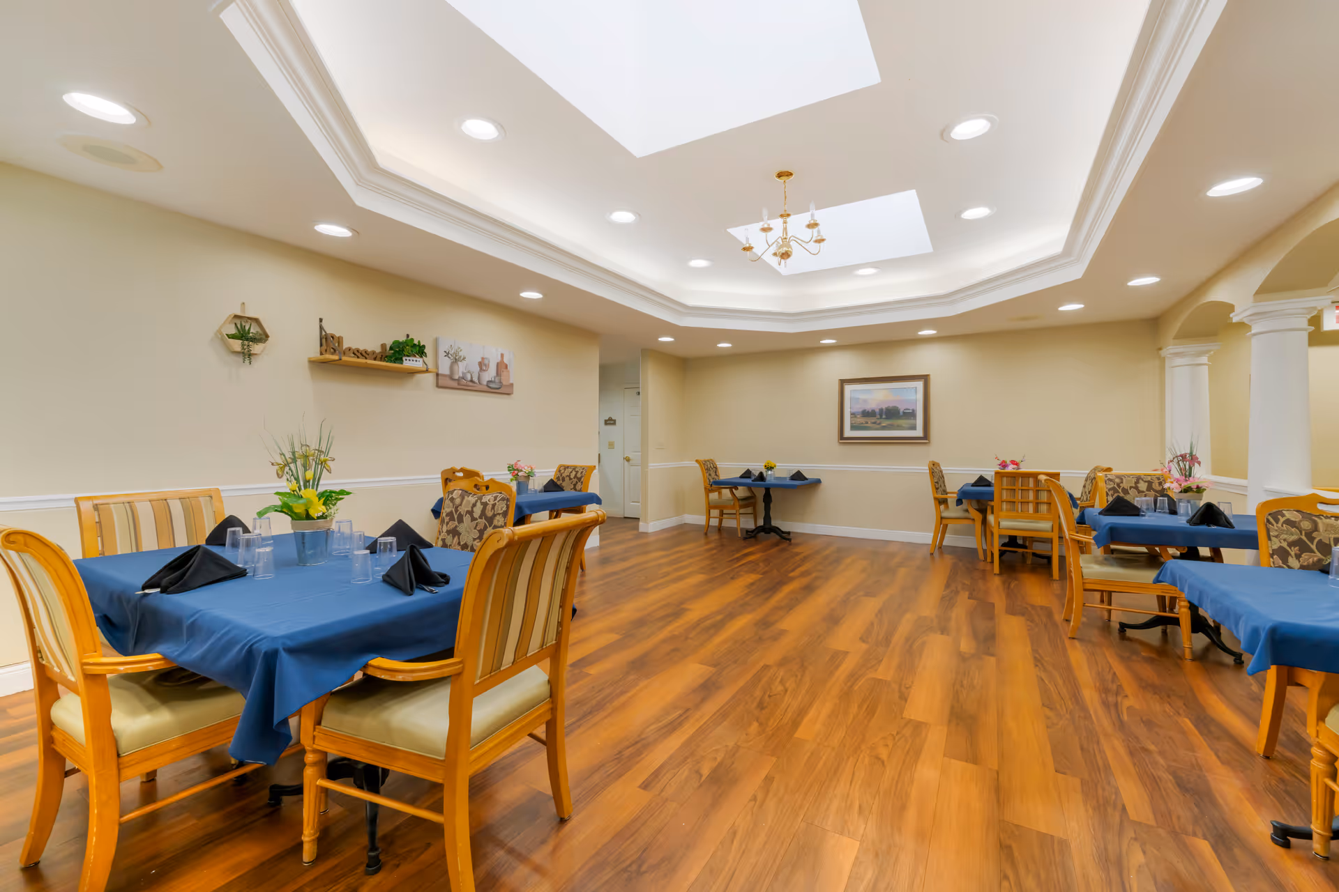 A dining room with several tables covered in blue tablecloths, each set with black napkins and clear drinking glasses. The room has wooden flooring, beige walls, recessed ceiling lights, and two skylights. There are decorative plants and framed artwork on the walls.