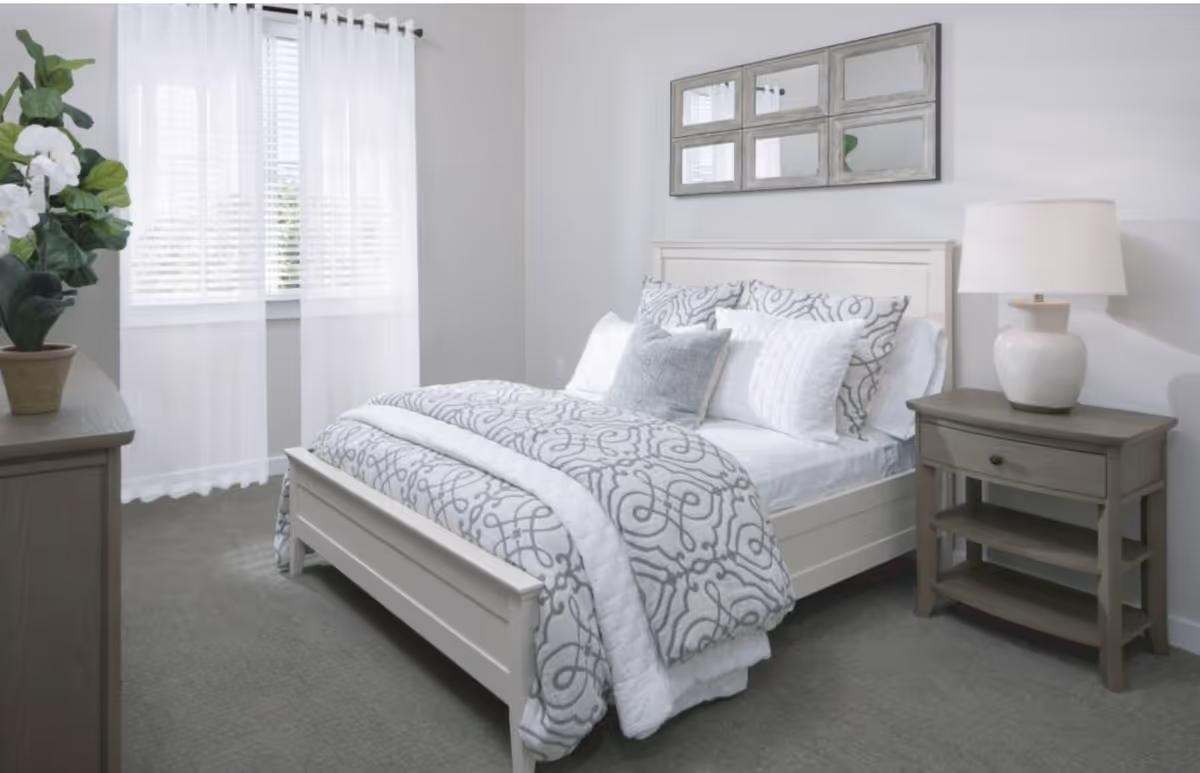 A bright bedroom with a white bed frame and a bed made with white and gray patterned bedding and multiple pillows. There is a wooden nightstand with a white lamp on the right side of the bed and a dresser with a potted plant on the left. A window with sheer white curtains allows natural light to fill the room. Above the bed is a decorative wall mirror with six panels.