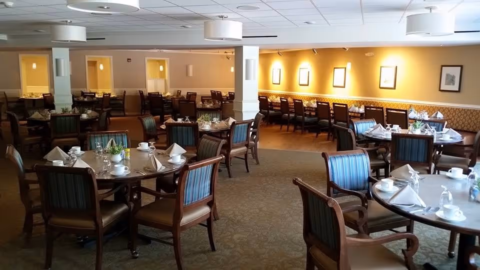 A spacious dining room with multiple round and rectangular tables set with white napkins, cups, glasses, and silverware. The room features wooden chairs with blue and green striped upholstery, warm yellow walls with framed artwork, and soft overhead lighting.