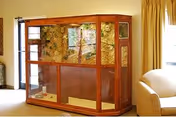 A large wooden and glass terrarium or display case situated in a living room area with beige walls and curtains, and a cream-colored armchair partially visible on the right side.