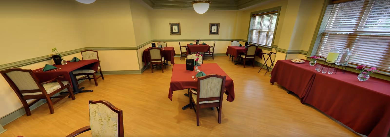 A dining room with several tables covered in red tablecloths, each set with green napkins, menus, and small flower vases. There is a side table near the window with a red tablecloth holding a beverage dispenser, glasses, and plates of cookies. The room has wooden floors, light yellow walls with green trim, and large windows with blinds.