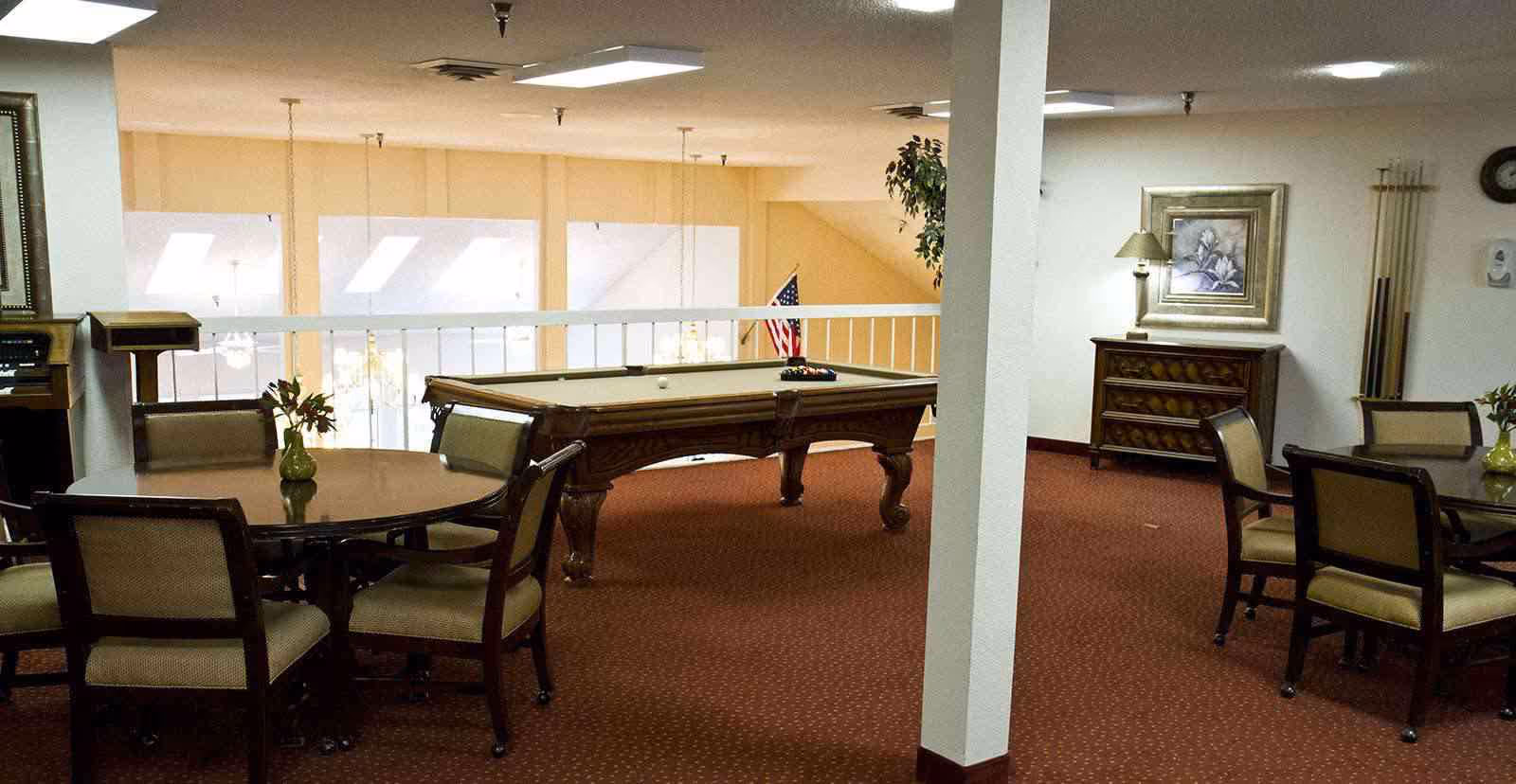 Interior view of a senior living facility common area with a pool table in the center, round tables with chairs around the room, a small cabinet with a lamp and framed artwork on the wall, and a rack holding pool cues. The room has carpeted flooring and a white railing overlooking a lower level.