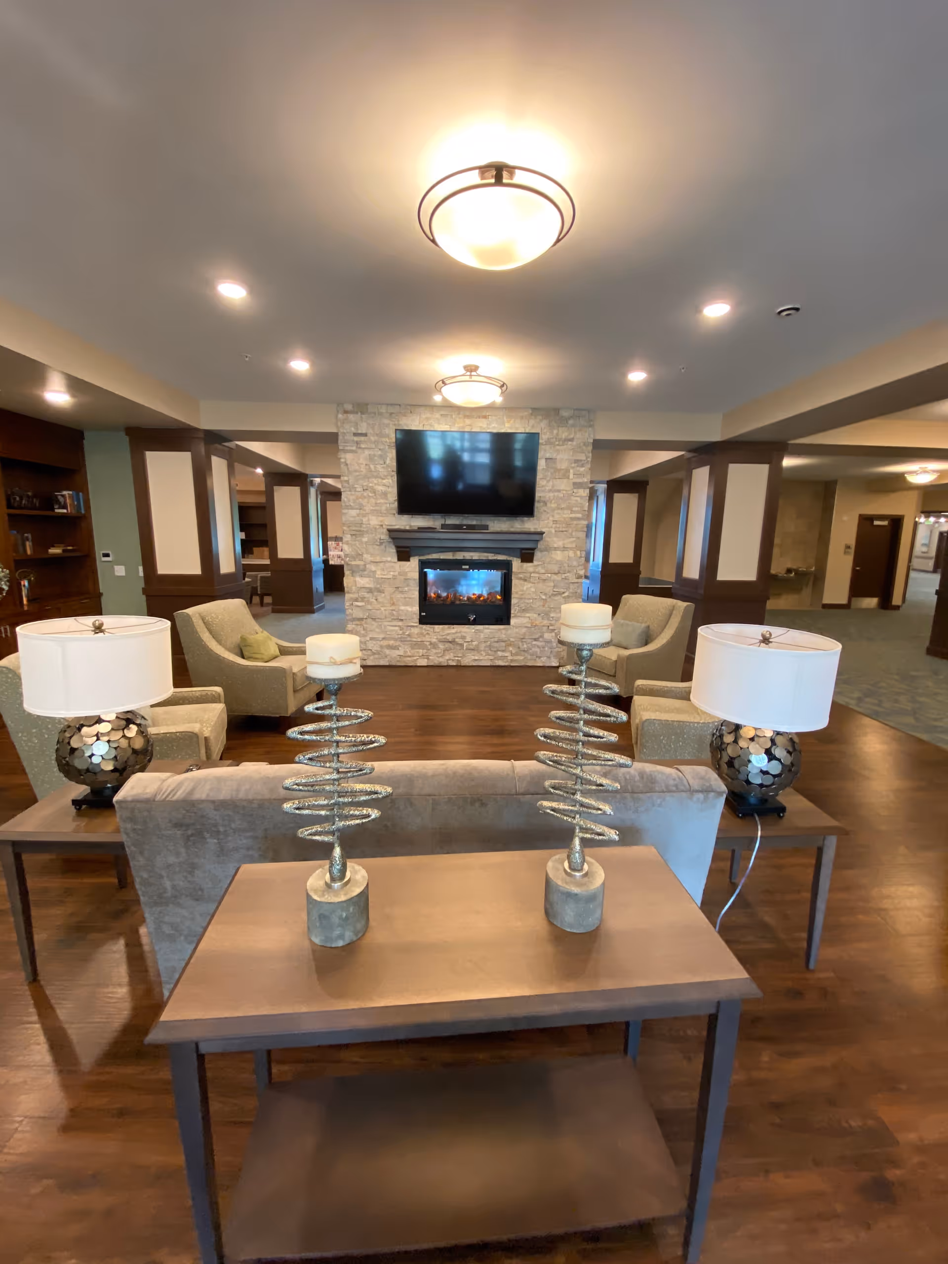 A cozy living room area with a stone fireplace and a mounted flat-screen TV above it. The room features four beige armchairs arranged around the fireplace, two side tables with decorative lamps, and a wooden console table with two spiral candle holders. The floor is wooden, and the ceiling has recessed lighting and two round ceiling lights.