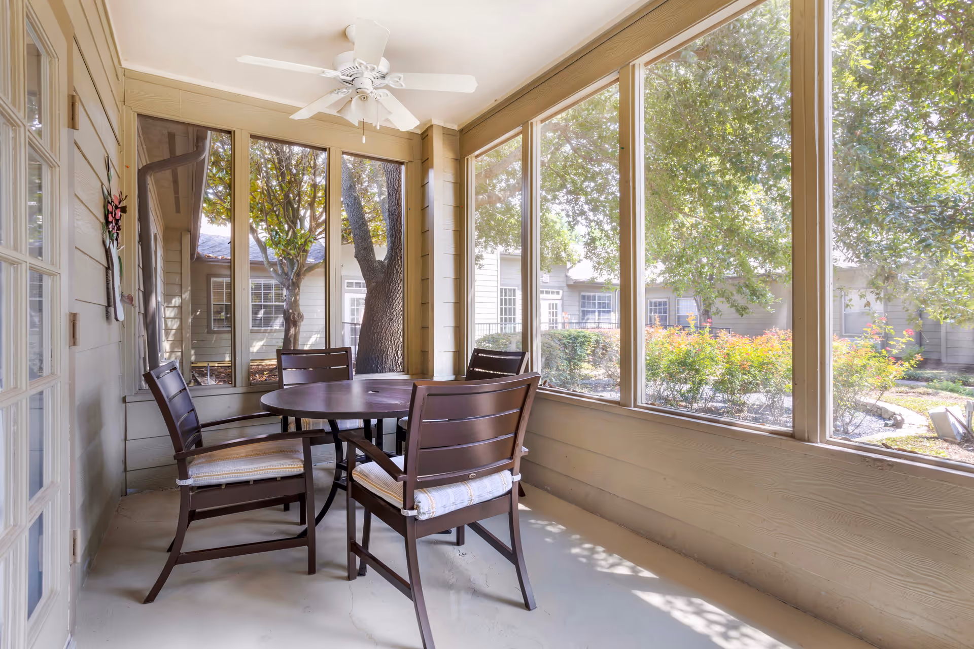 A screened-in porch area with a round table and four cushioned chairs. The porch has large windows providing a view of trees, bushes, and a building exterior. A ceiling fan is mounted above the table.