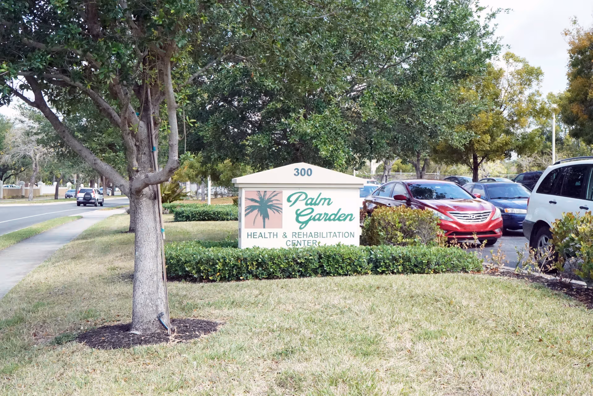 Outdoor view of the Palm Garden Health & Rehabilitation Center sign with the number 300 on top, surrounded by green bushes and trees, with parked cars and a road visible in the background.