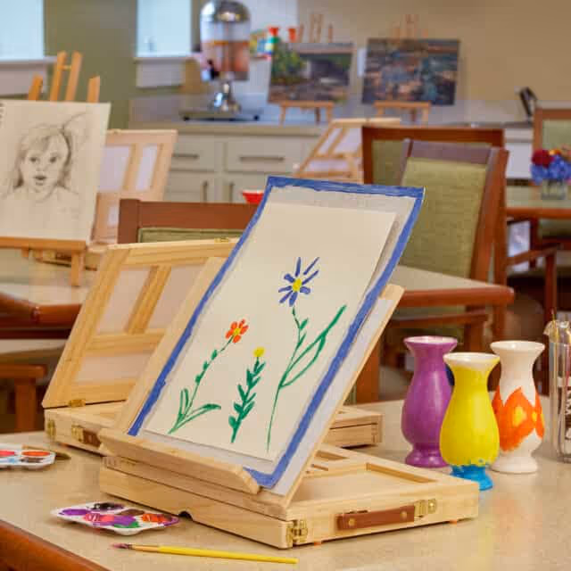 A table in a senior living facility art room with a wooden easel holding a painting of colorful flowers. In the background, there are other paintings, chairs, and art supplies visible.