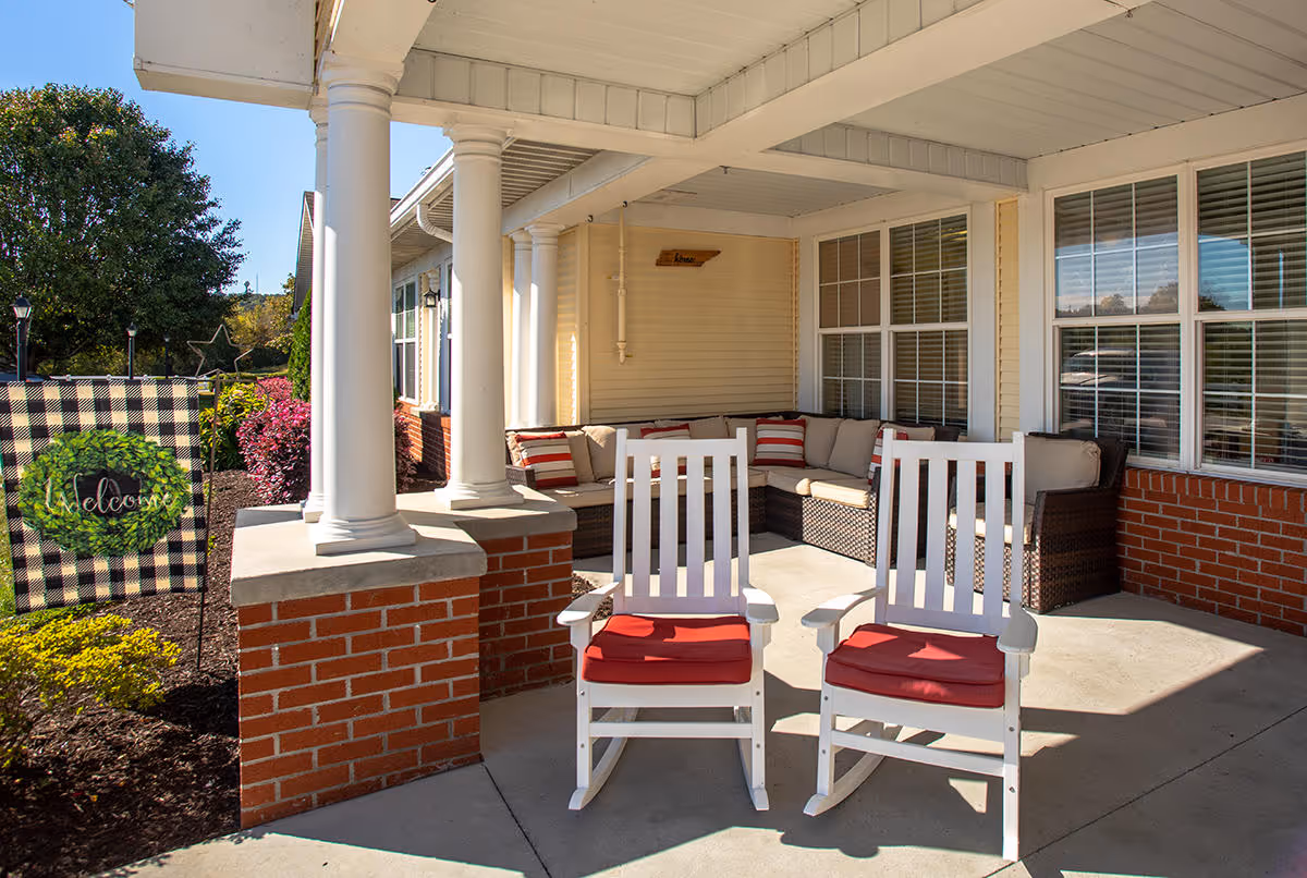 Outdoor covered patio area with two white rocking chairs with red cushions in the foreground, a wicker sectional sofa with beige cushions and red-striped pillows in the background, white columns, brick half-walls, and a welcome flag with a green wreath design on the left side.