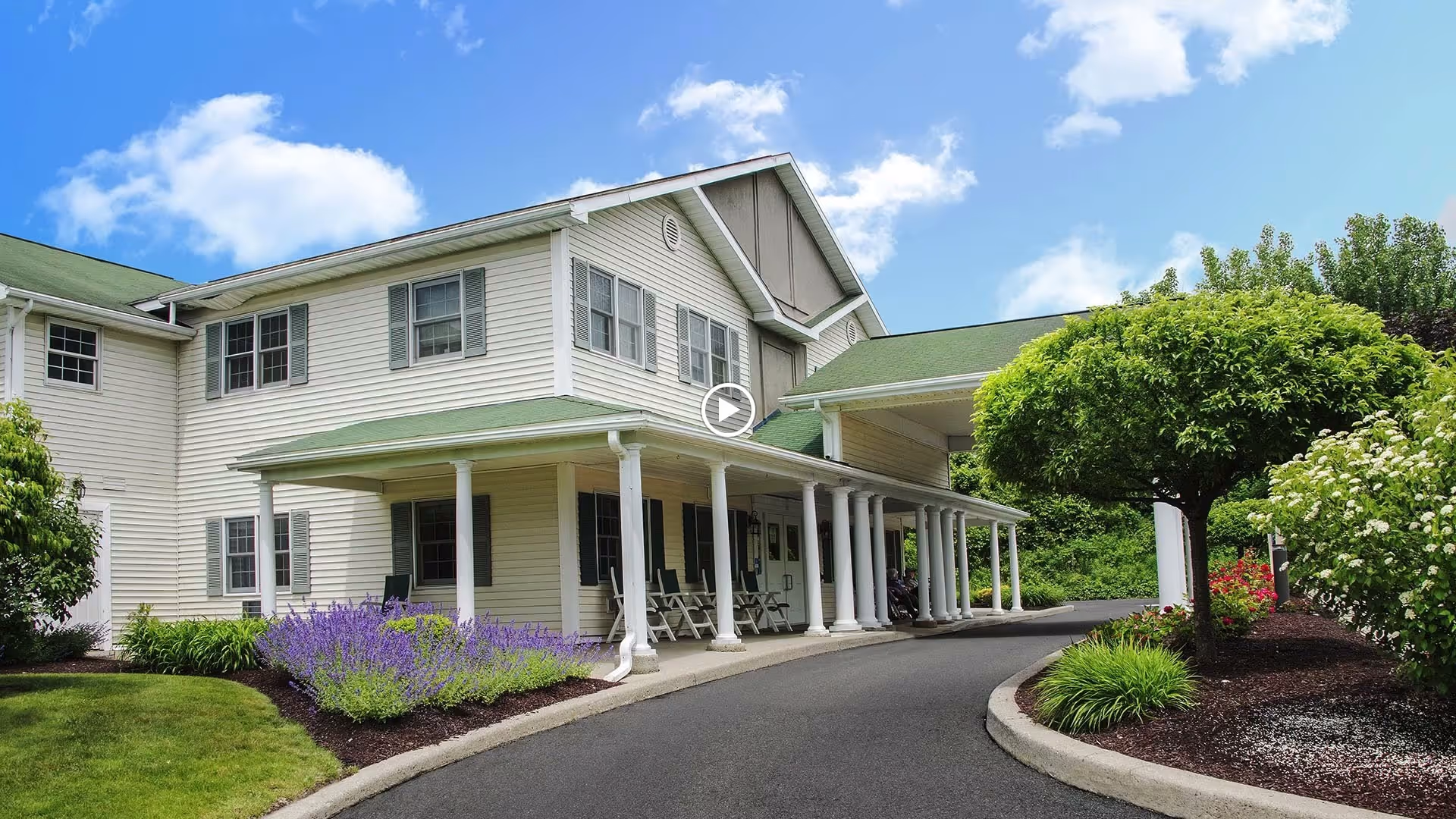 Exterior view of a two-story senior living facility building with white siding and green roof. The building features a covered entrance supported by white columns, surrounded by well-maintained landscaping including bushes, flowering plants, and a small tree. The sky is blue with some clouds.