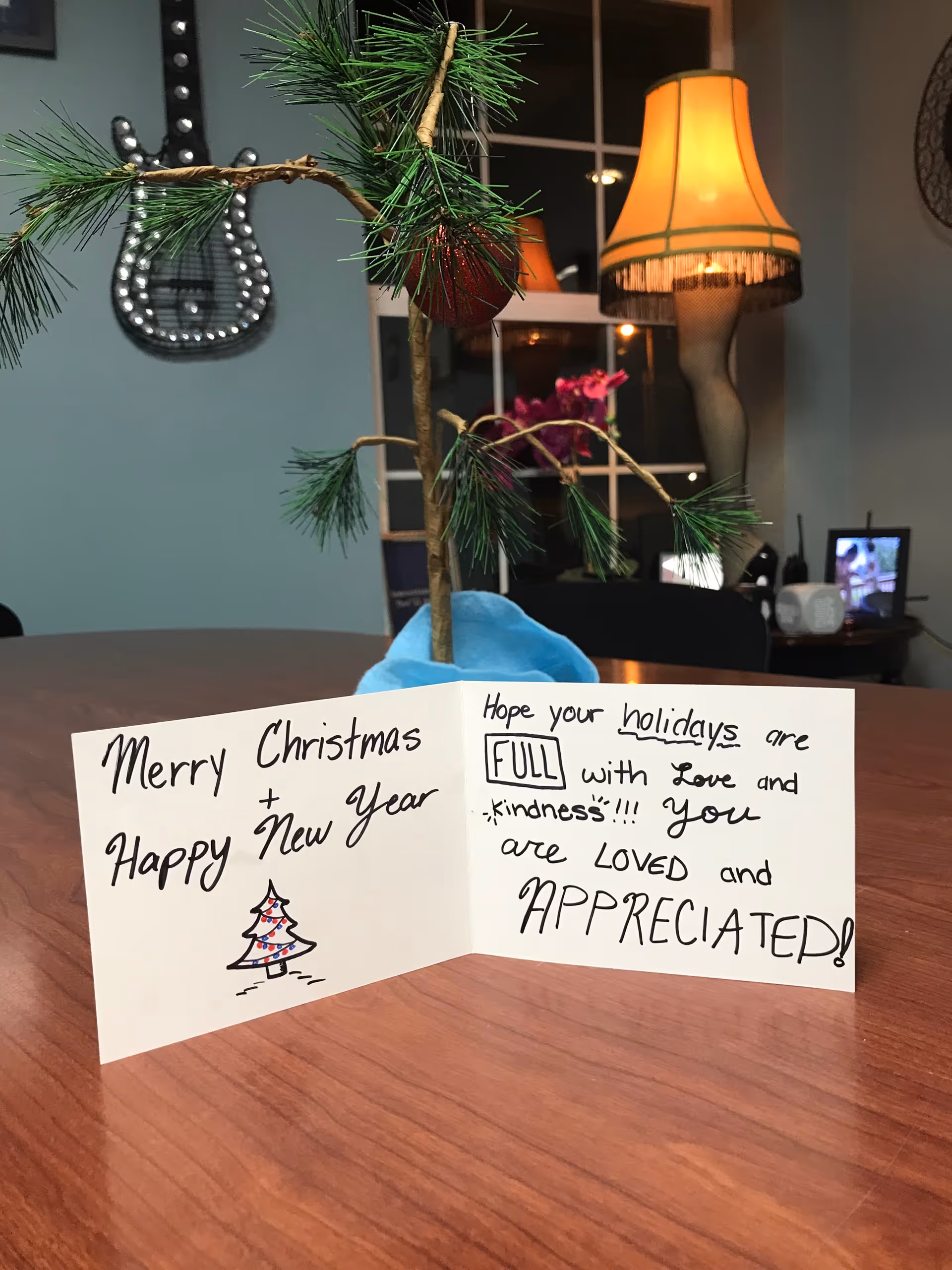 A small, sparse Christmas tree with a single red ornament sits on a wooden table. In front of the tree is a handwritten holiday card that reads, 'Merry Christmas + Happy New Year' with a small drawing of a Christmas tree, and 'Hope your holidays are FULL with love and kindness!!! You are LOVED and APPRECIATED!' In the background, there is a leg lamp, a window, and some furniture in a cozy room.